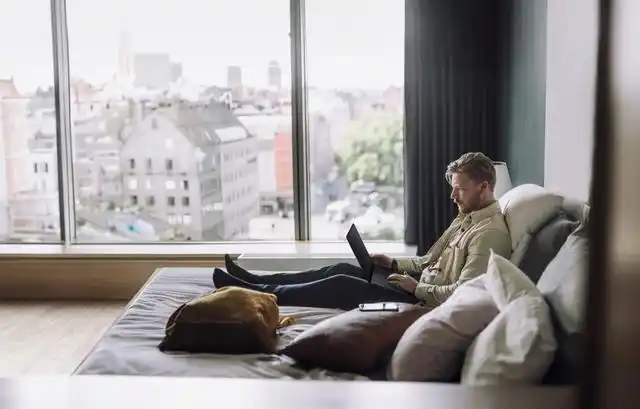 Man working on a laptop while sitting on a bed in a modern room with a large window and city view