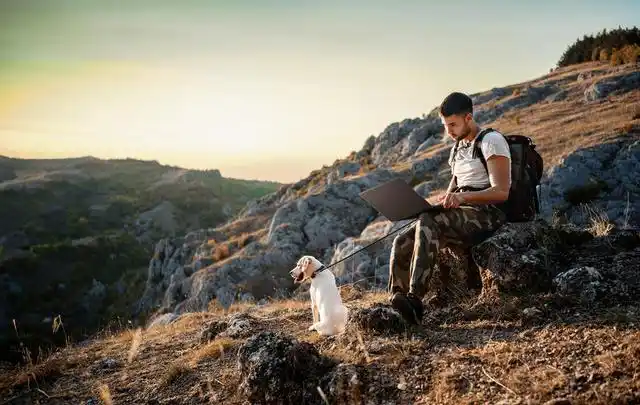 Man using a laptop outdoors while sitting on a rocky hilltop with a small dog on a leash beside him during sunset