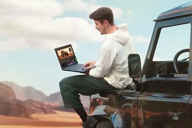 A man sits on the hood of a black off-road vehicle in a desert landscape, working on a laptop. He is wearing a white hoodie and dark pants, with mountains and a partly cloudy sky in the background