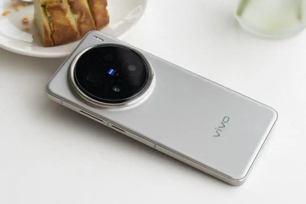 Vivo X300 smartphone in silver color with a prominent circular Zeiss camera module placed on a white table beside a dessert plate