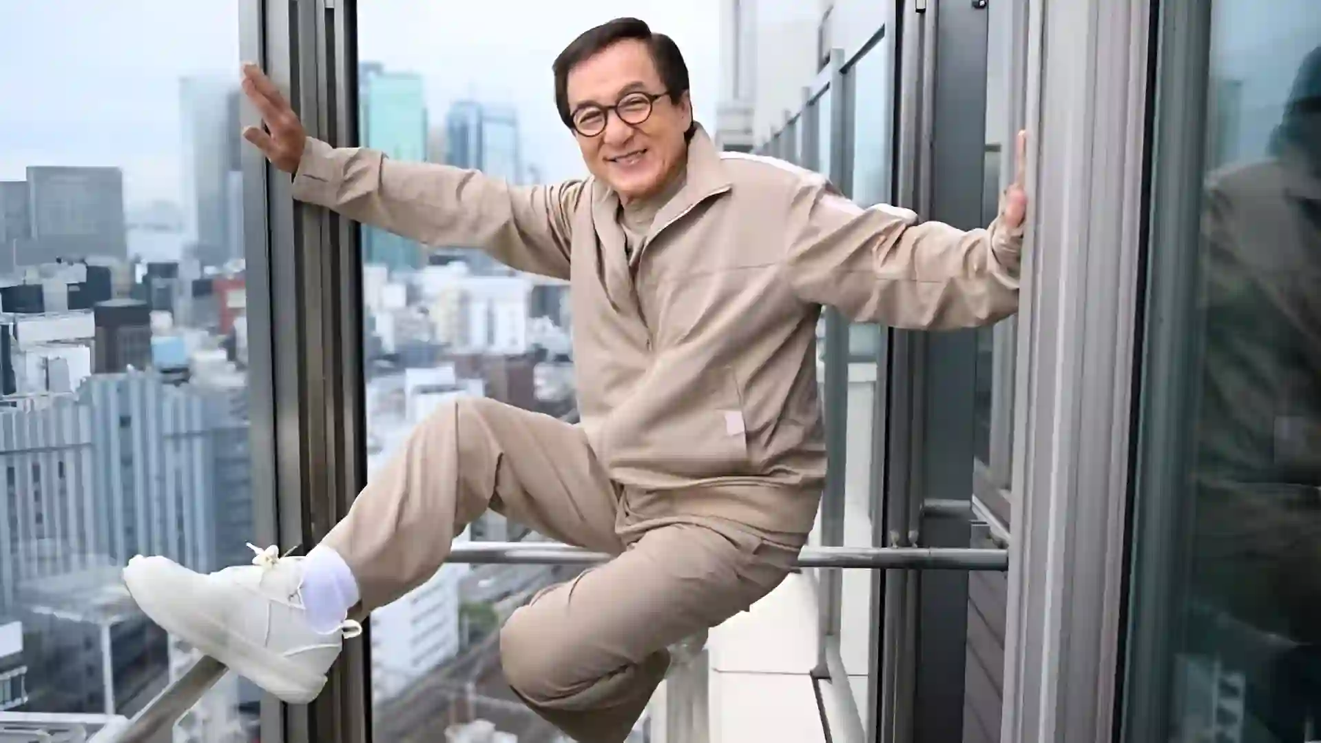 Jackie Chan, smiling and wearing a tan jumpsuit, doing a classic stunt pose on a high-rise balcony with a city view