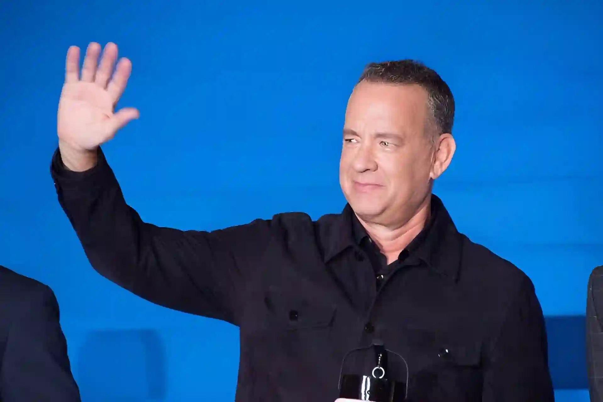 Tom Hanks smiling and waving at an event, wearing a black shirt with a blue background