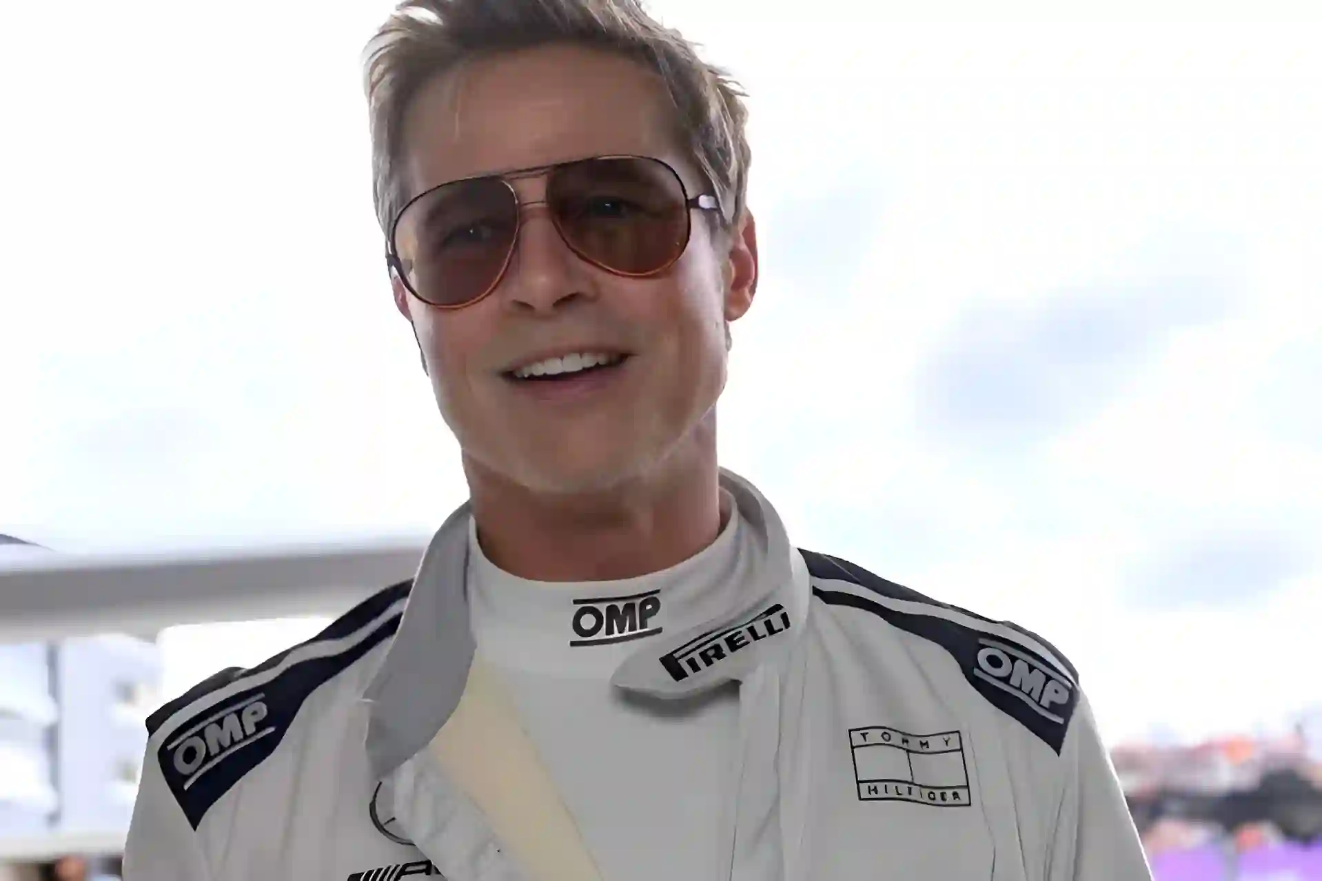 Brad Pitt smiling, wearing a white OMP racing suit and sunglasses