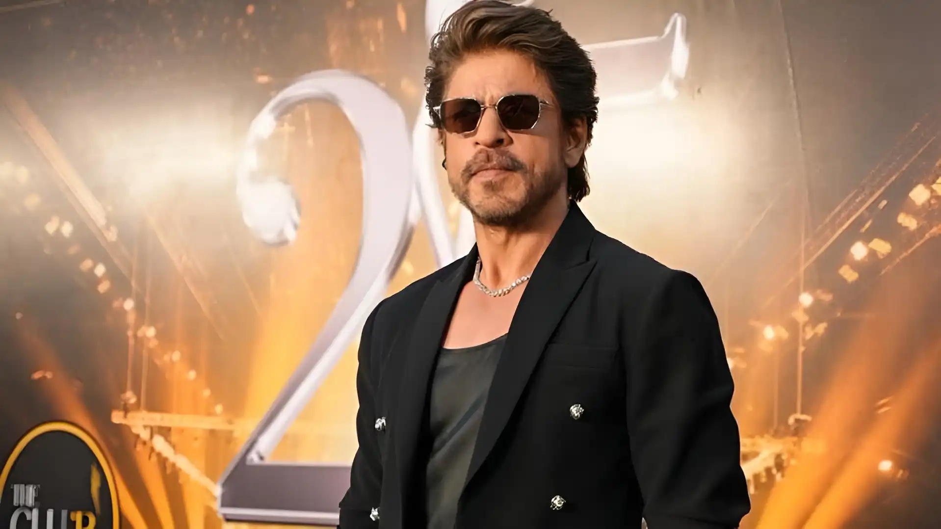 Shah Rukh Khan wearing sunglasses and a black suit, standing in front of a shimmering golden background