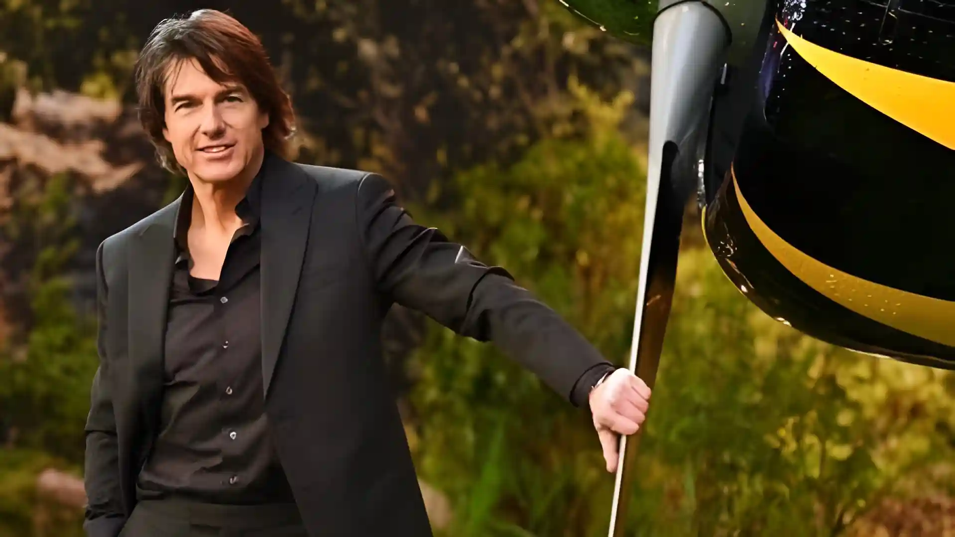Tom Cruise smiling, leaning against the propeller of a plane, with a blurred outdoor background
