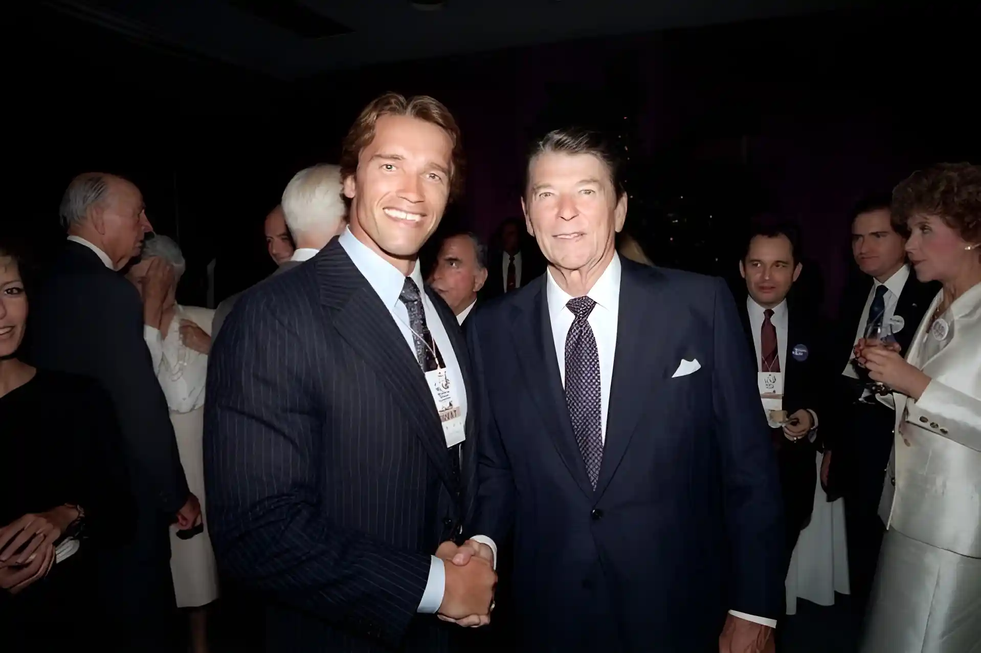 Arnold Schwarzenegger shaking hands with Ronald Reagan at an event, with other people visible in the background.