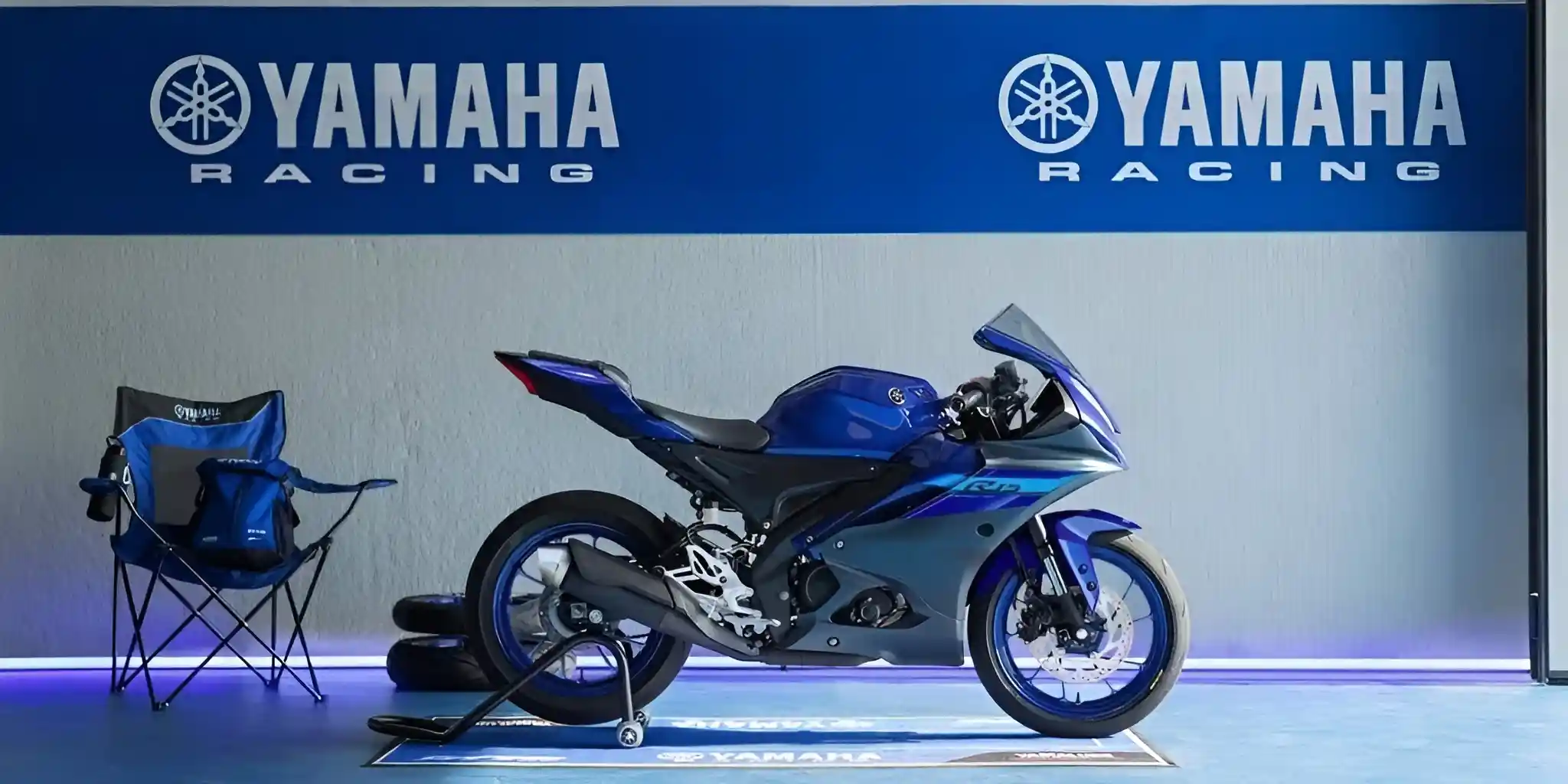 A dark blue and gray Yamaha R15M motorcycle in a garage with Yamaha Racing branding, next to a folding chair.