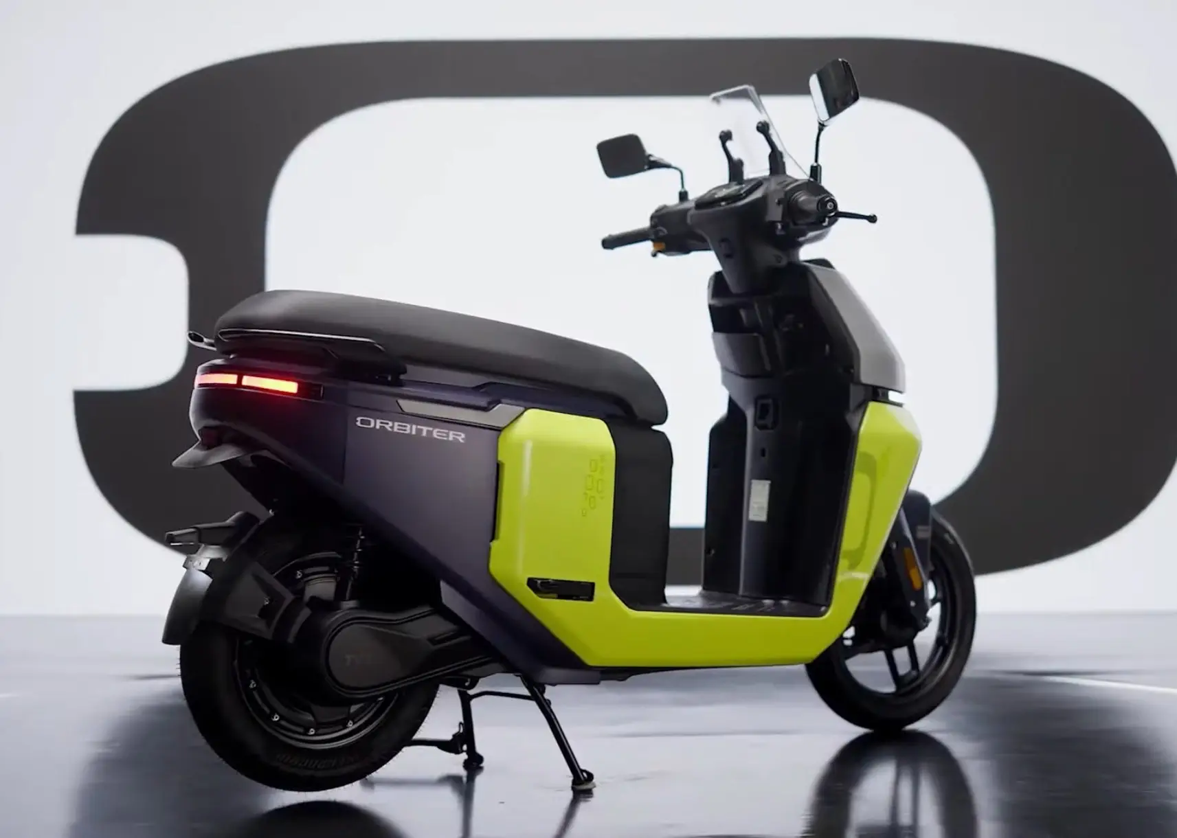 A yellow and a brown TVS Orbiter electric scooter on display on a stage. The TVS
Orbiter logo is above the scooters.