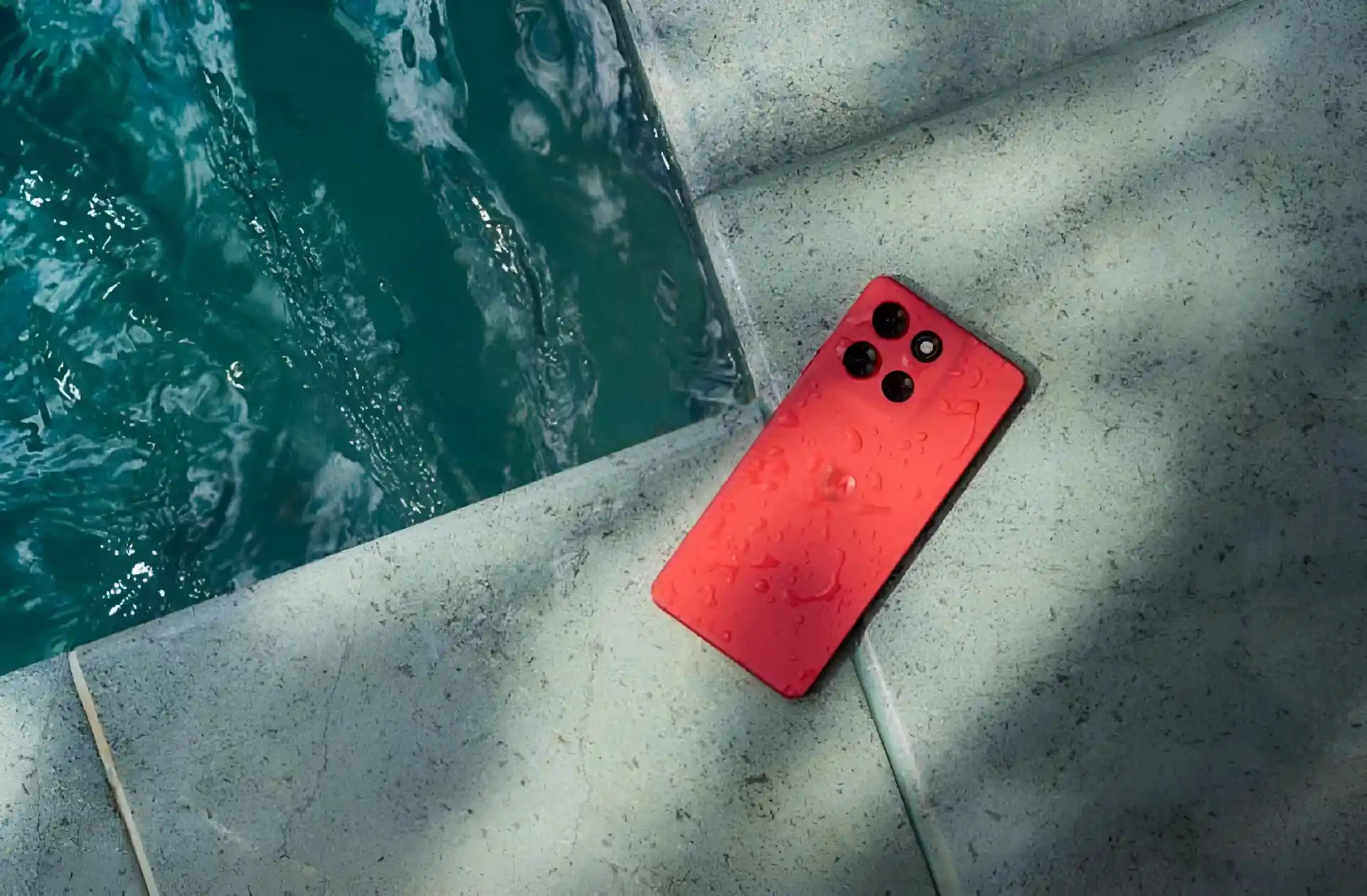 A red Motorola Moto G86 5G phone, with water droplets on its back, rests on the edge of a swimming pool.